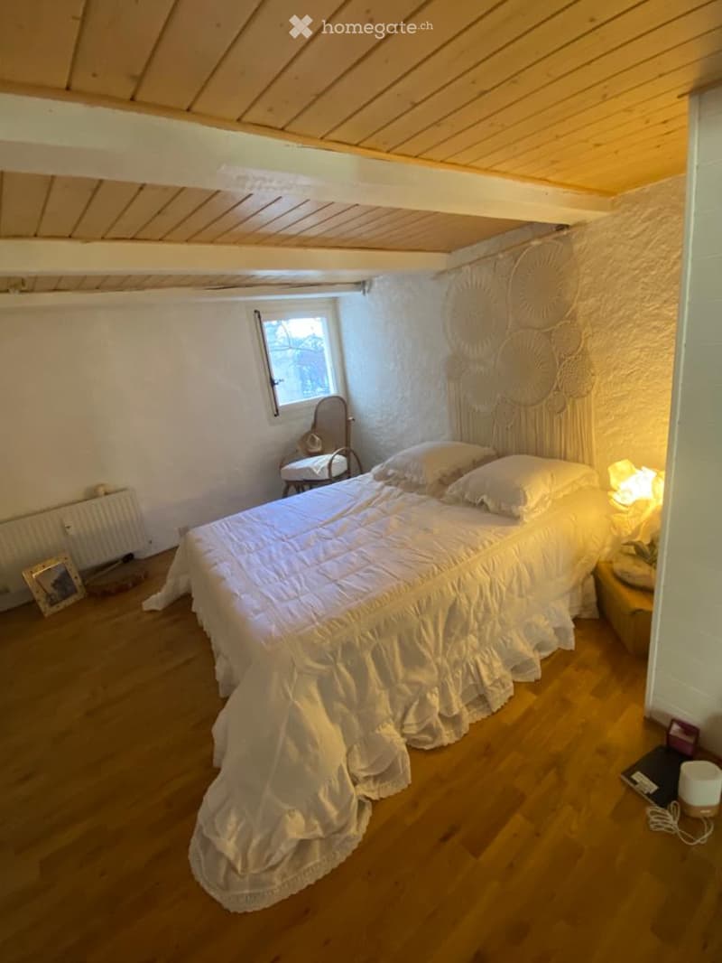 Cozy attic bedroom with wooden floors, slanted wooden ceiling, white walls, a large bed with white bedding, a nightstand with a lamp, a small chair, and a decorative wall hanging.