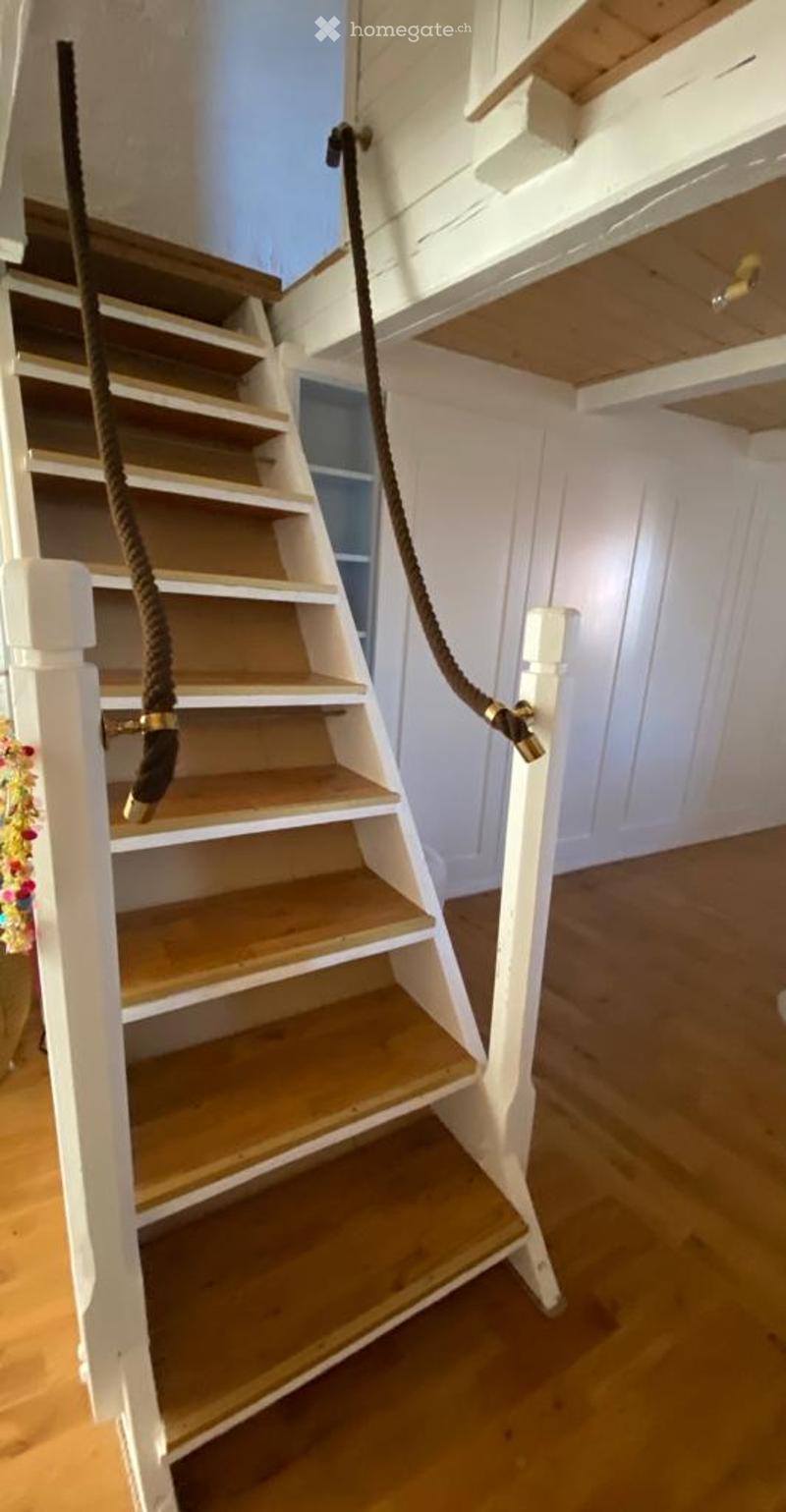 Wooden staircase with white railings and rope handrails, leading to an upper level with exposed wooden beams on the ceiling.