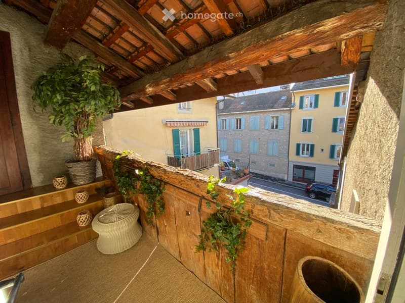 Wooden balcony with tiled floor, wicker chair, potted plants, and view of the street and neighboring buildings.