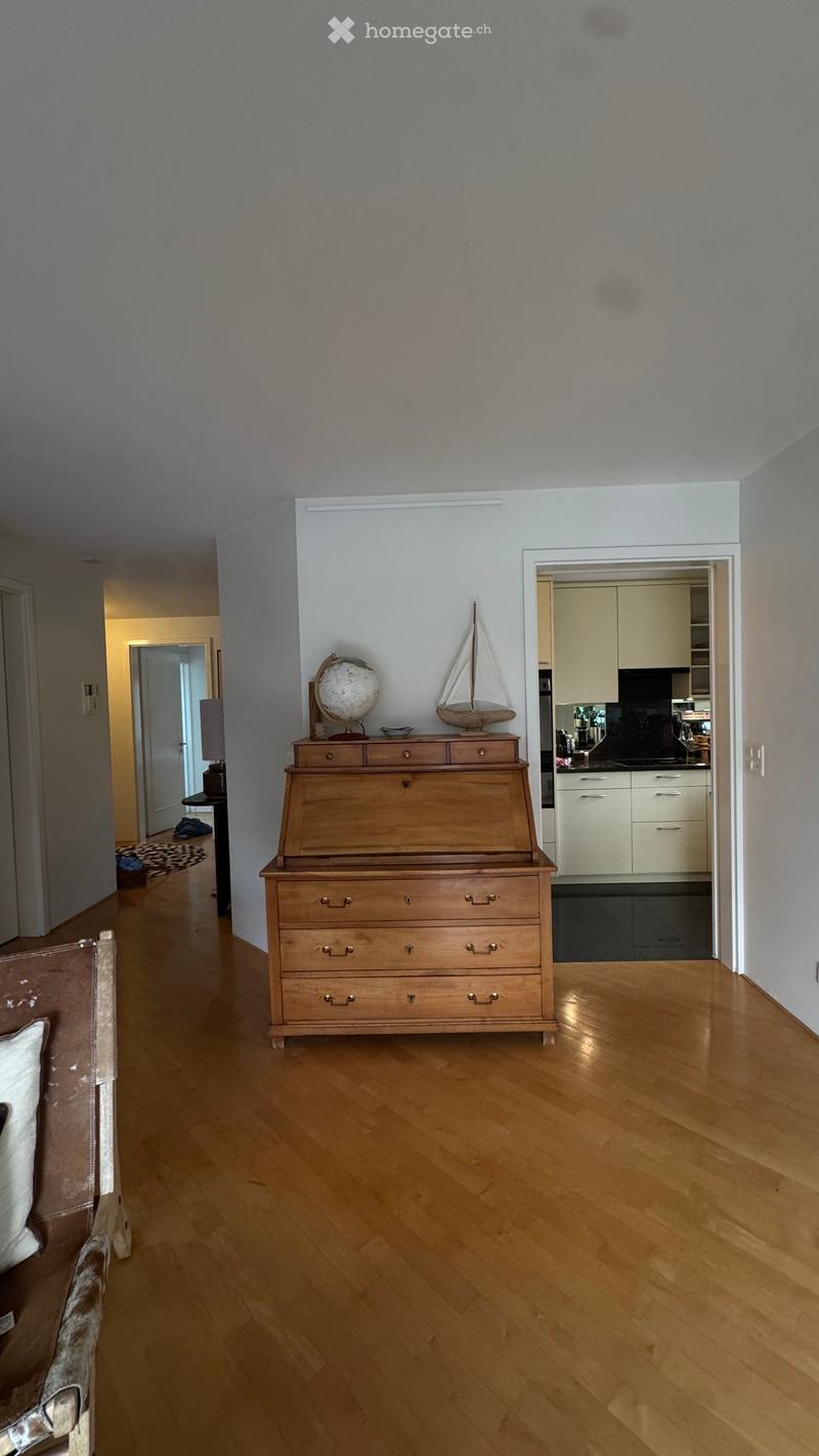 A well-lit hallway with wooden flooring, a chest of drawers with a globe and model sailboat on top, and a glimpse of a modern kitchen with white cabinets and stainless steel appliances.