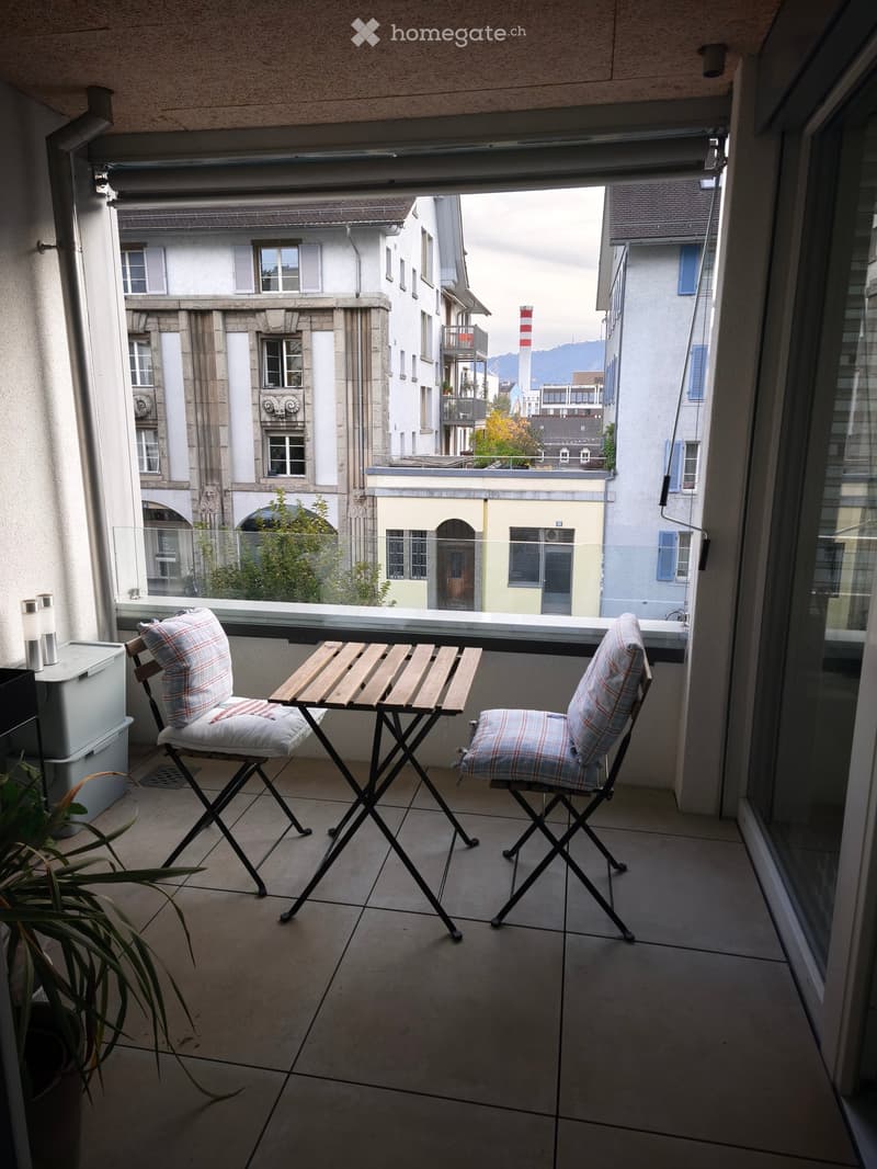 Balcony with a small wooden table and two matching chairs, overlooking a cityscape with buildings and a distant view of mountains.