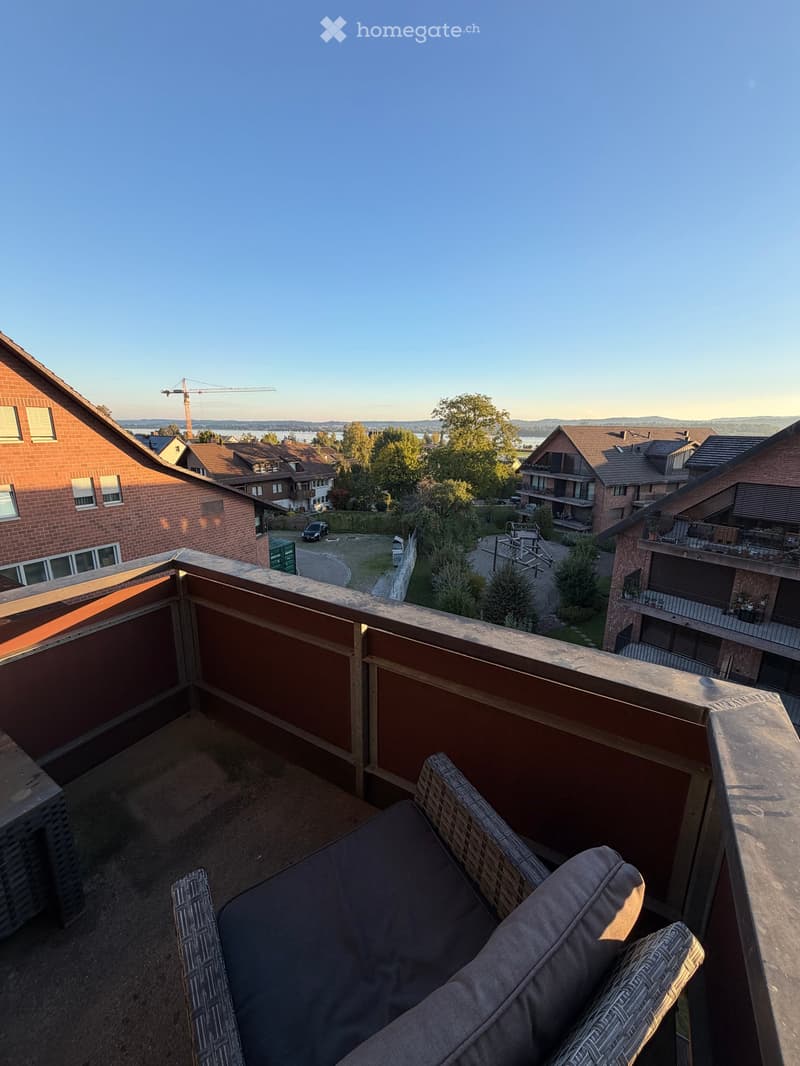 Balcony with a wooden railing, a grey cushioned sofa, and a view of a residential area with houses, trees, and a distant body of water.