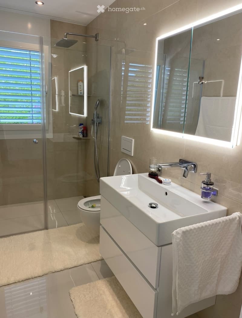 Modern bathroom with a white sink, mirror with LED lighting, toilet, glass-enclosed shower, and towel on a rack.