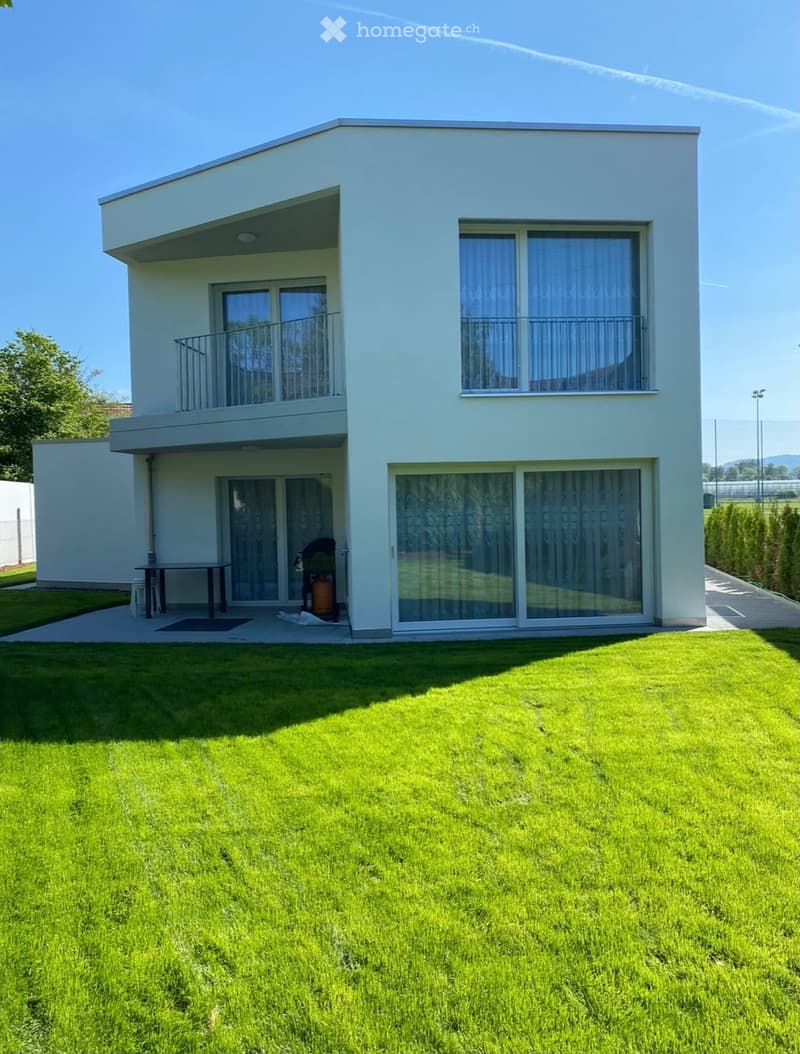 Modern two-story house with light blue exterior, large windows, and a balcony on the upper floor. The house features a well-maintained lawn in the front yard and a small patio area with a table and chairs near the entrance.