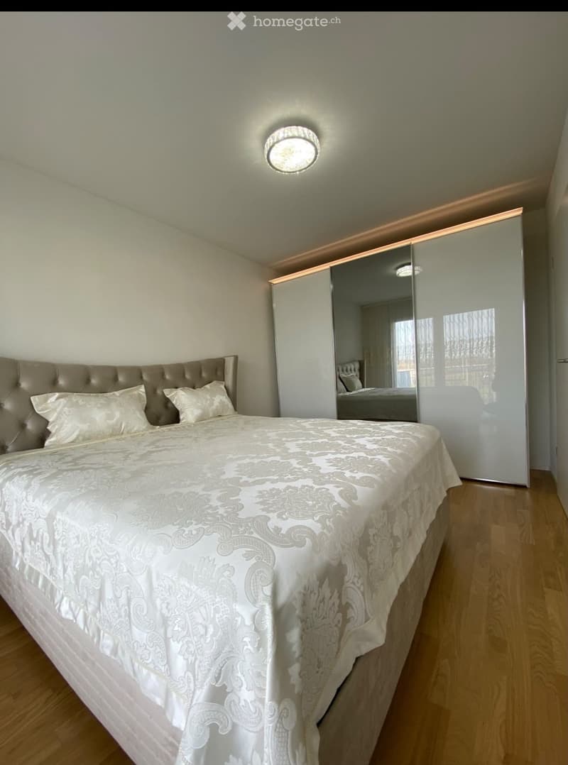Elegant bedroom with a large, tufted headboard bed, white bedding, and a mirrored wardrobe. The room features wooden flooring and modern lighting.