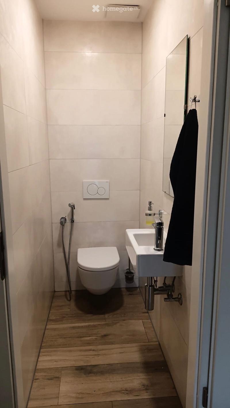 Modern, compact bathroom with white tiled walls, wooden floor, toilet, sink with modern faucet, mirror, and a black towel hanging on the wall.