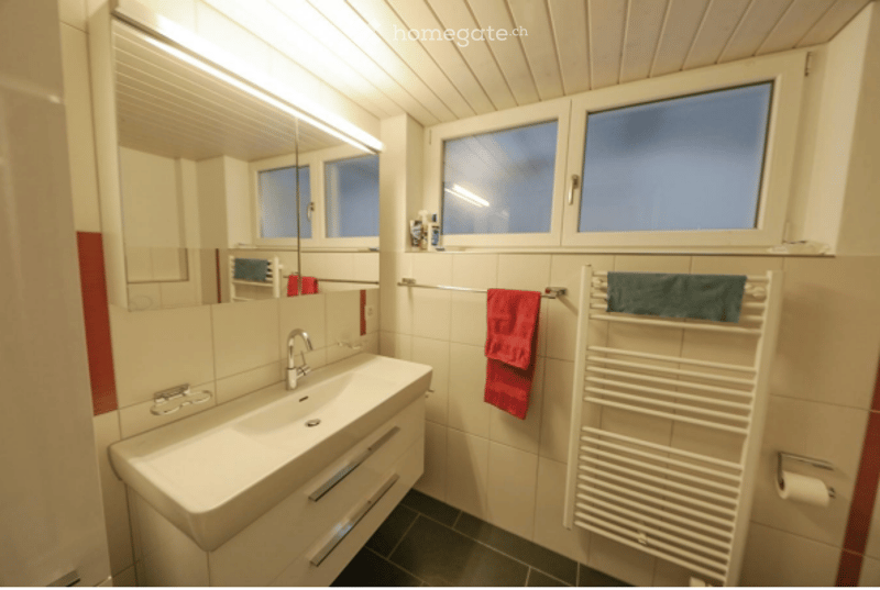 Modern bathroom with white tiled walls, a large sink with a mirror, a towel rack with towels, and a radiator.