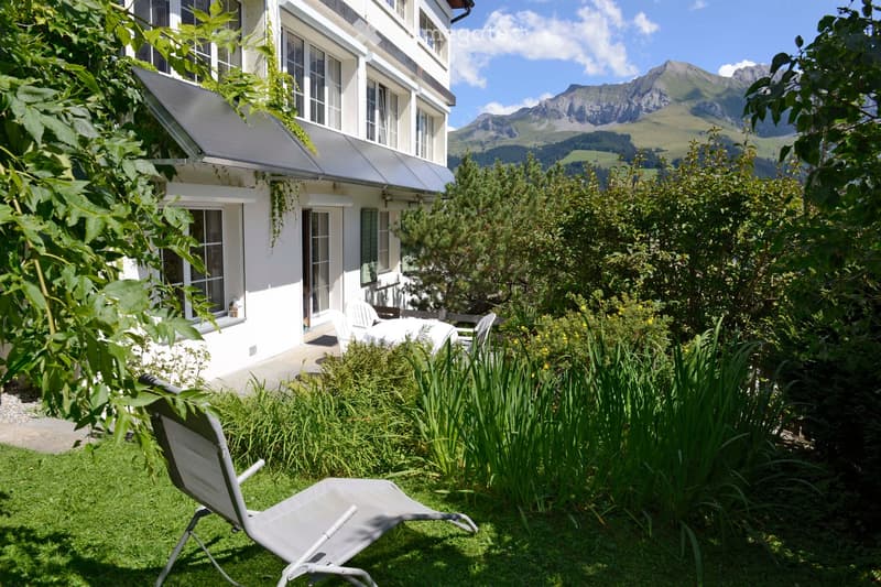 A well-maintained garden area with a lounge chair, surrounded by lush greenery and a view of a multi-story white building with large windows and a covered patio. The background features a scenic mountain range under a clear blue sky.