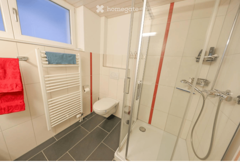 Modern bathroom with white tiles, a glass-enclosed shower, a toilet, a towel rack with towels, and a window above the sink.