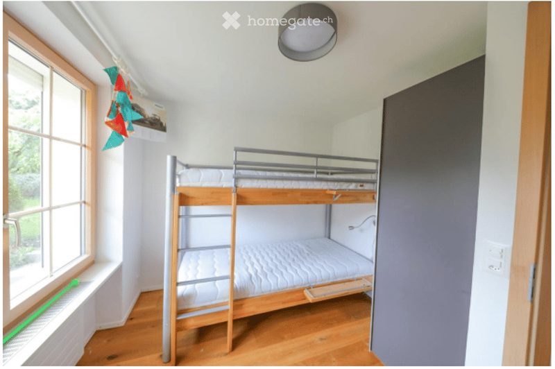 A small bedroom with a wooden bunk bed, white walls, wooden flooring, and a window with a view of greenery.
