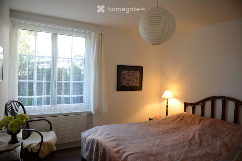 Cozy bedroom with a double bed, a wooden chair, a small table with a vase of flowers, a bedside lamp, and a large window with white curtains. A decorative picture frame hangs on the wall, and a large paper lantern hangs from the ceiling.