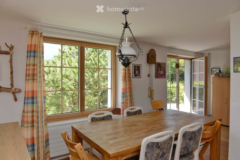 Bright kitchen with large windows, wooden dining table surrounded by chairs, cabinet, picture frames, and a hanging light fixture.