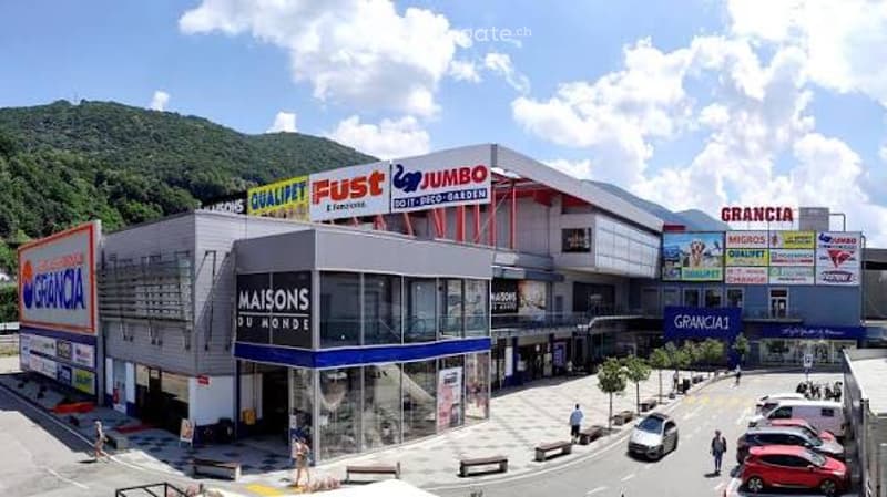 Large commercial building with multiple storefronts, including 'JUMBO' and 'MAISONS DU MONDE', surrounded by parking area and greenery.