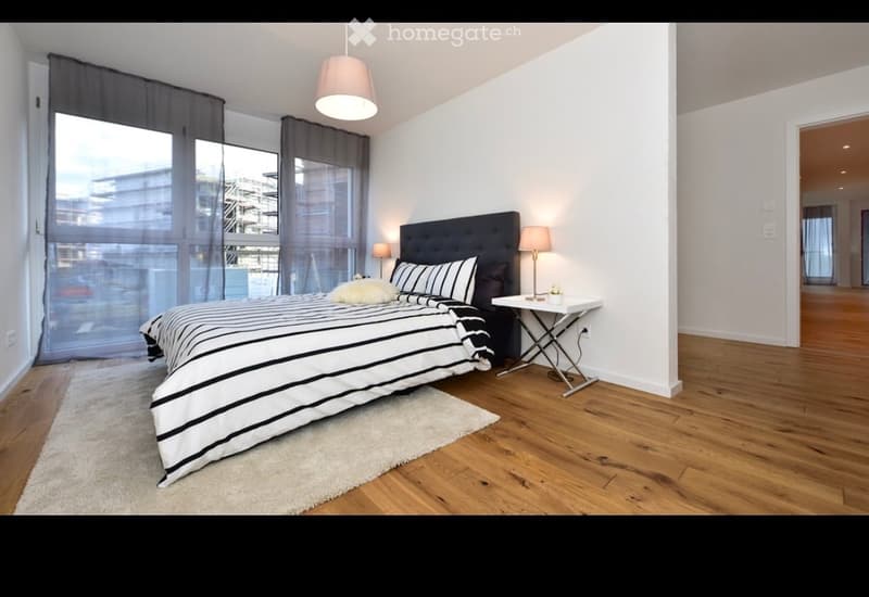 Modern bedroom with a large bed, nightstand, table lamp, and large windows with curtains. The room features wooden flooring and a white rug.