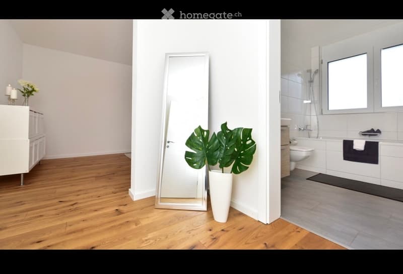Modern, minimalist room with wooden flooring, large mirror, potted plant, and adjacent bathroom with bathtub, toilet, and shower.