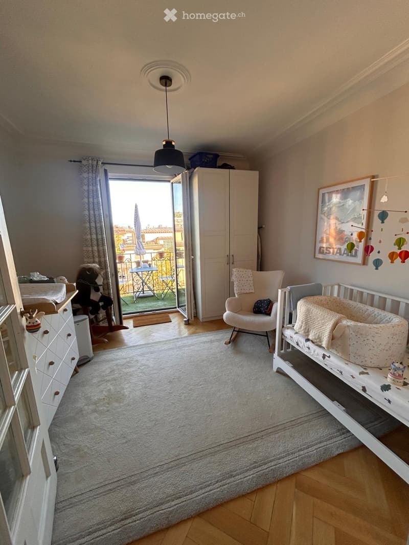 Brightly lit bedroom with a crib, rocking chair, cabinet, and toys. Large window opens to a balcony with outdoor furniture.