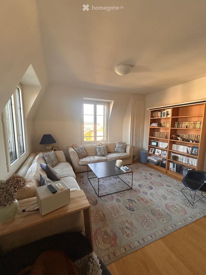 Bright living room with wooden floors, large windows, a sectional sofa, coffee table, bookshelf, and a patterned rug.