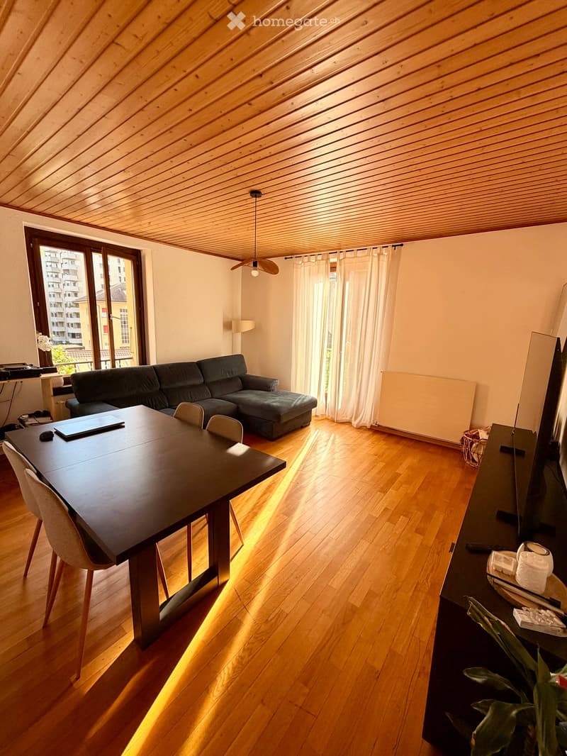 Bright living room with wooden floors, a grey sectional sofa, a dark dining table with chairs, a television, and a ceiling fan. Large windows with white curtains let in natural light.