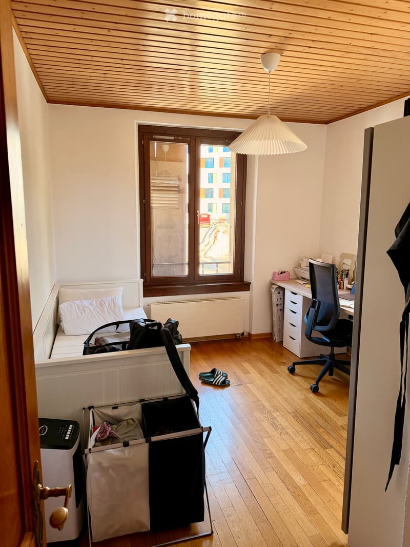A small, well-lit room with a wooden floor, a white sofa, a desk with a chair, a lamp, a printer, and a baby stroller. The room has a large window with blinds and a view of the street.