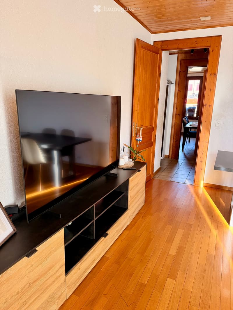 A modern living room with wooden flooring, a large flat-screen TV on a wooden stand, and a hallway leading to another room with a dining table and chairs.