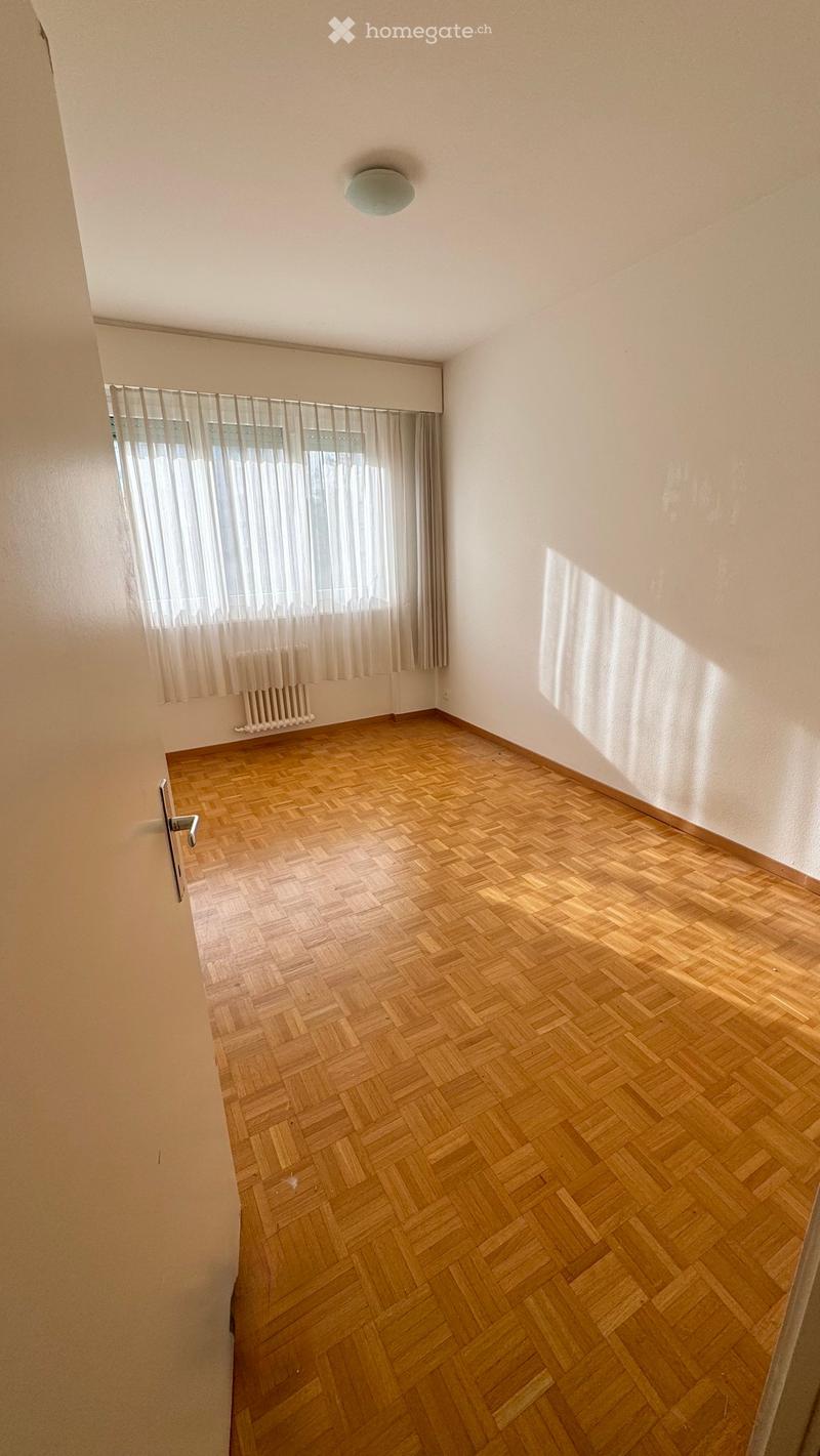 Empty room with wooden herringbone flooring, white walls, a window with sheer curtains, and a radiator below the window.