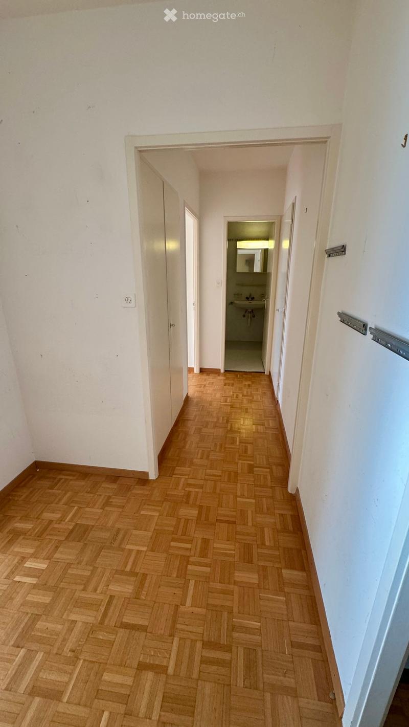 Empty hallway with wooden herringbone flooring, white walls, and doors leading to other rooms, including a visible bathroom.