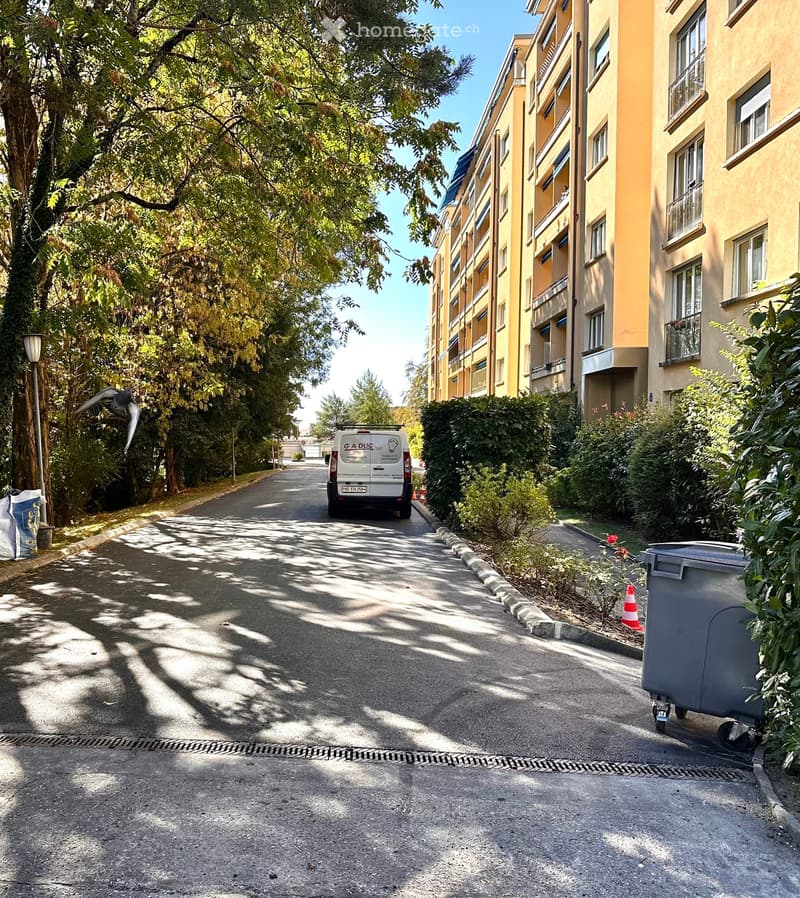A tree-lined residential street with a multi-story building on the right, featuring balconies and windows, and a parked van in the distance.