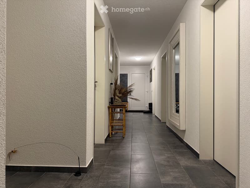 A long, narrow hallway with tiled flooring and white walls, featuring a small wooden table with a plant and a statue on it, and a mirror on the right wall.