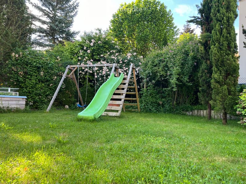 Lush garden with a green slide, swing, and climbing frame surrounded by trees and flowering bushes.