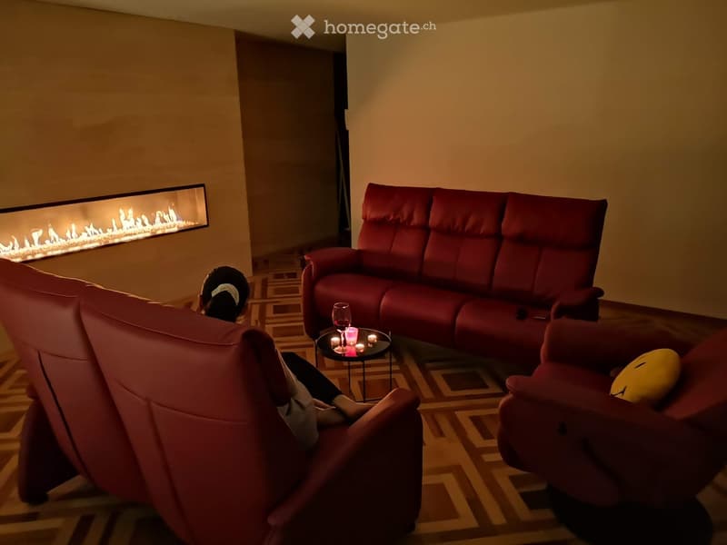 Modern living room with red leather sofa, armchair, small table with wine glass and candles, and a fireplace with a simulated flame display.