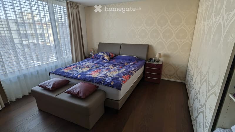 Cozy bedroom with a large bed, patterned duvet, two pillows, a nightstand with a lamp, and a small bench at the foot of the bed.
