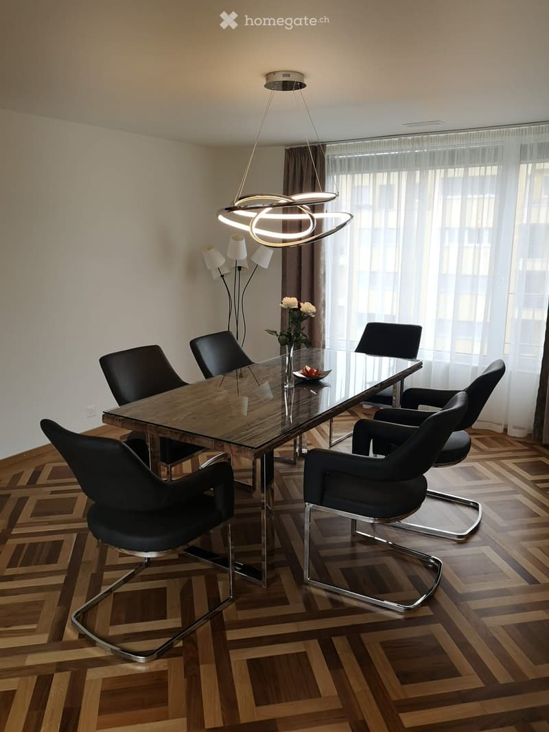Modern dining room with a glass-top wooden dining table, six black chairs, a pendant light fixture, and a wall-mounted lamp. The room features wooden herringbone flooring and large windows with sheer curtains.