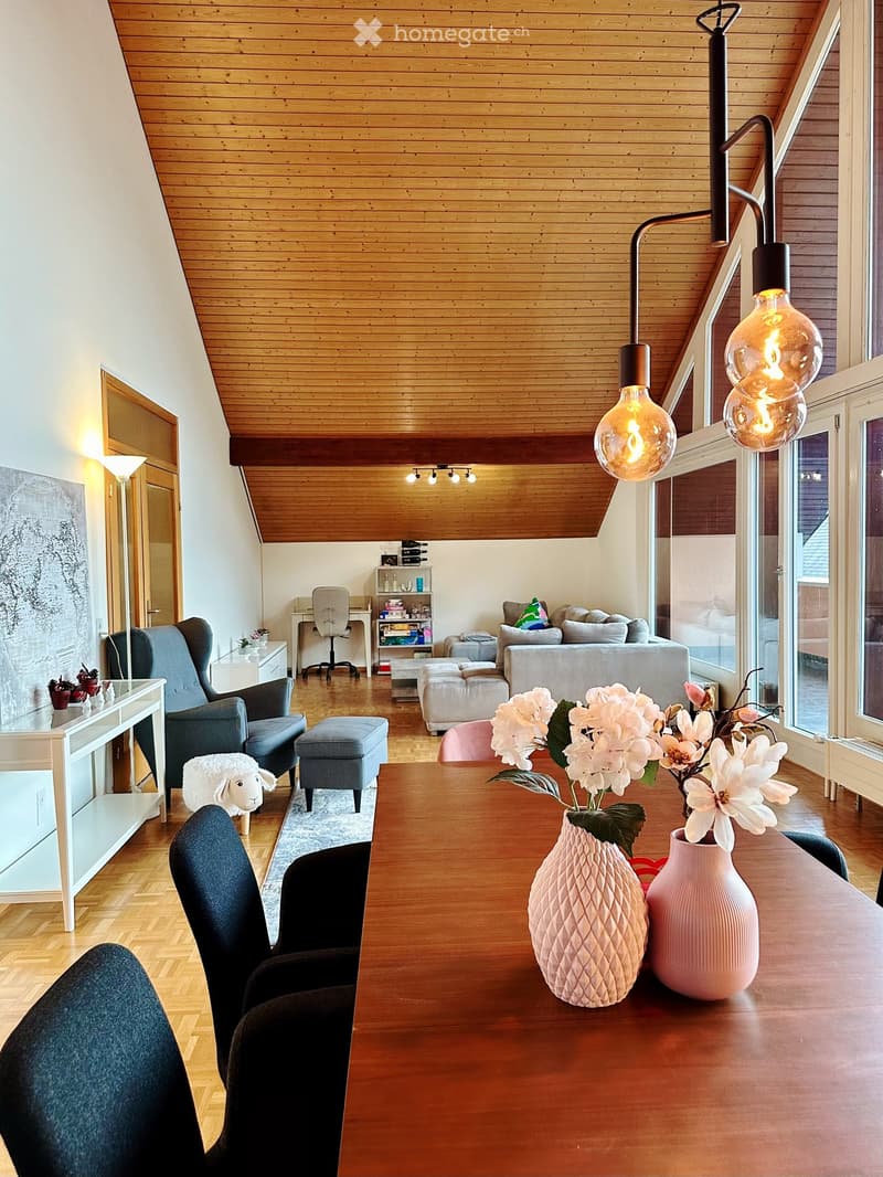 Bright, modern living room with wooden floors and a slanted wooden ceiling. Features a sofa, chairs, a coffee table with vases, a small shelf, and a hanging lamp with multiple bulbs.