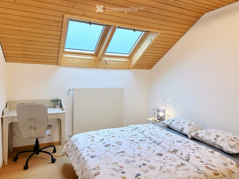 Cozy attic bedroom with wooden ceiling, skylight windows, a double bed with floral bedding, a desk with a chair, and a small side table with a lamp and plant.