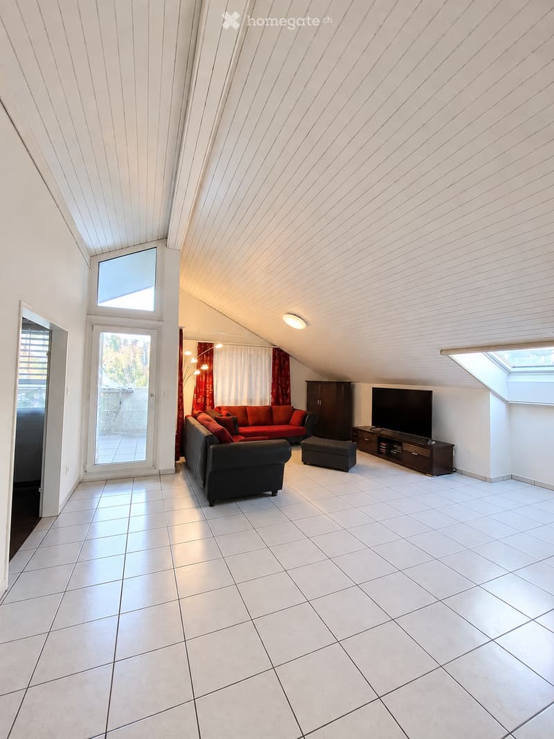 Modern living room with white tiled flooring, slanted ceiling, large windows, and a balcony. Features a black sofa, red ottoman, TV, and wooden cabinet.