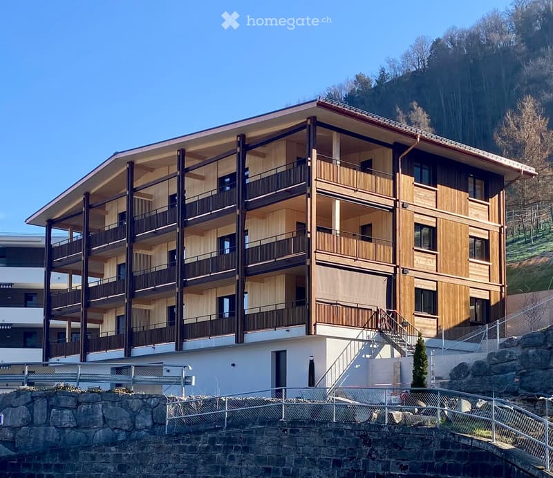 Modern multi-story building with wooden balconies and railings, situated on a raised ground floor with stone steps leading up.