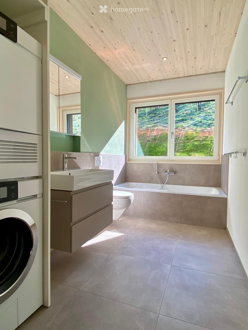 Modern bathroom with a large window offering a view of greenery, featuring a sink, bathtub, toilet, and washing machine.