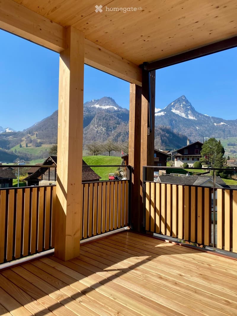 Wooden balcony with a railing, overlooking a scenic mountain landscape with snow-capped peaks and a village below.