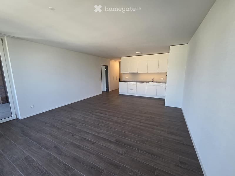 Empty room with dark wooden flooring and white walls, featuring a small white kitchen area in the background.