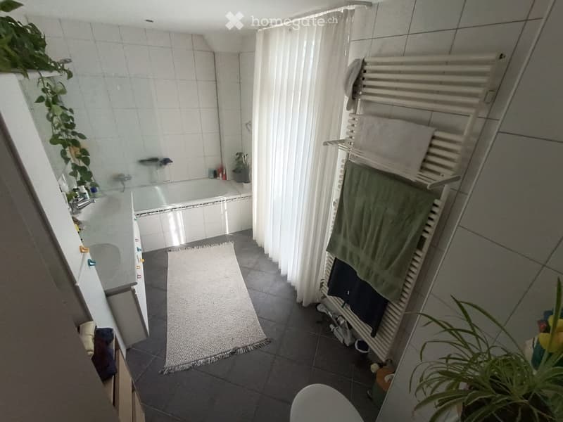Modern bathroom with a bathtub, sink, radiator, and towels. Features a white curtain, a small plant, and a grey rug on the floor.