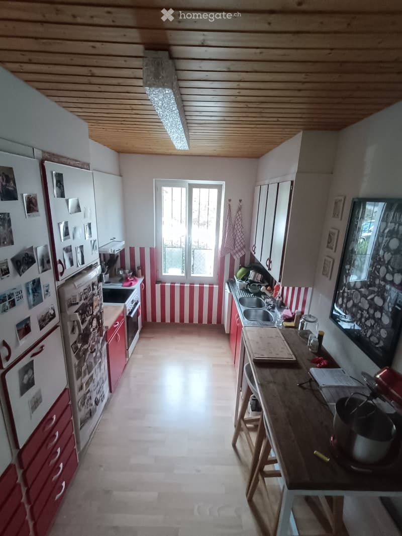 Bright kitchen with red and white striped backsplash, wooden ceiling, and natural light from a window. Features a refrigerator, oven, sink, counter, and wooden stool.