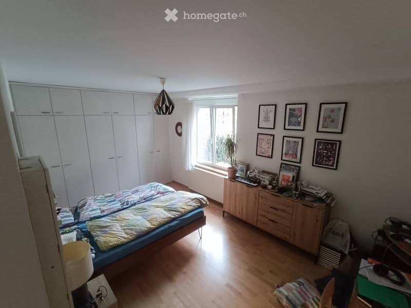 Bright bedroom with wooden flooring, large window, bed with colorful bedding, nightstand, chair, small table with keyboard and plant, framed pictures on wall, and built-in wardrobe.