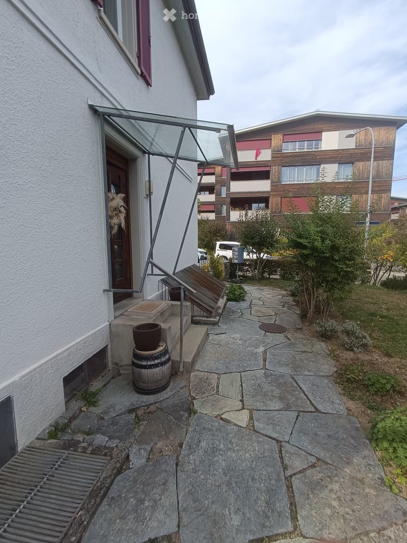 Stone-paved pathway leading to a house entrance with a glass canopy, wooden barrel, and small plants.