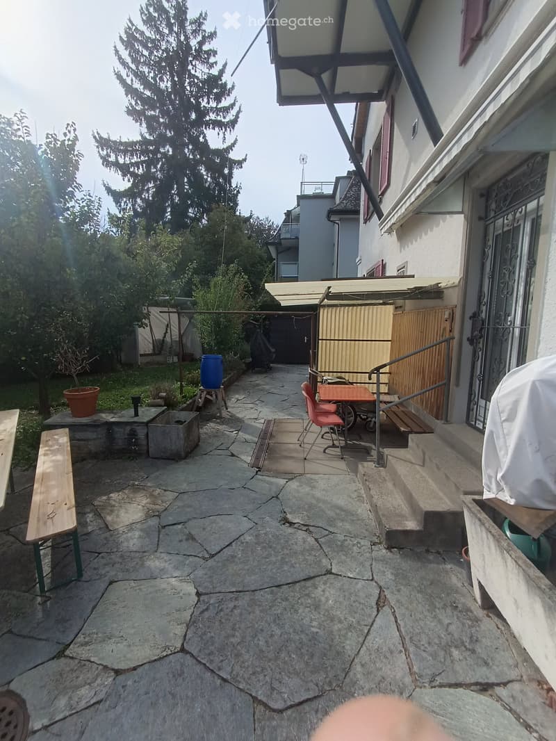 Outdoor patio area with stone pavement, wooden bench, small table with chairs, potted plants, and steps leading to a building entrance.