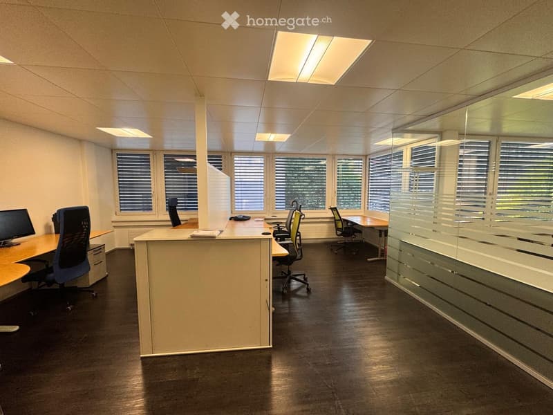 Office space with desks, chairs, computer monitors, and a telephone, featuring large windows with blinds and a glass partition.