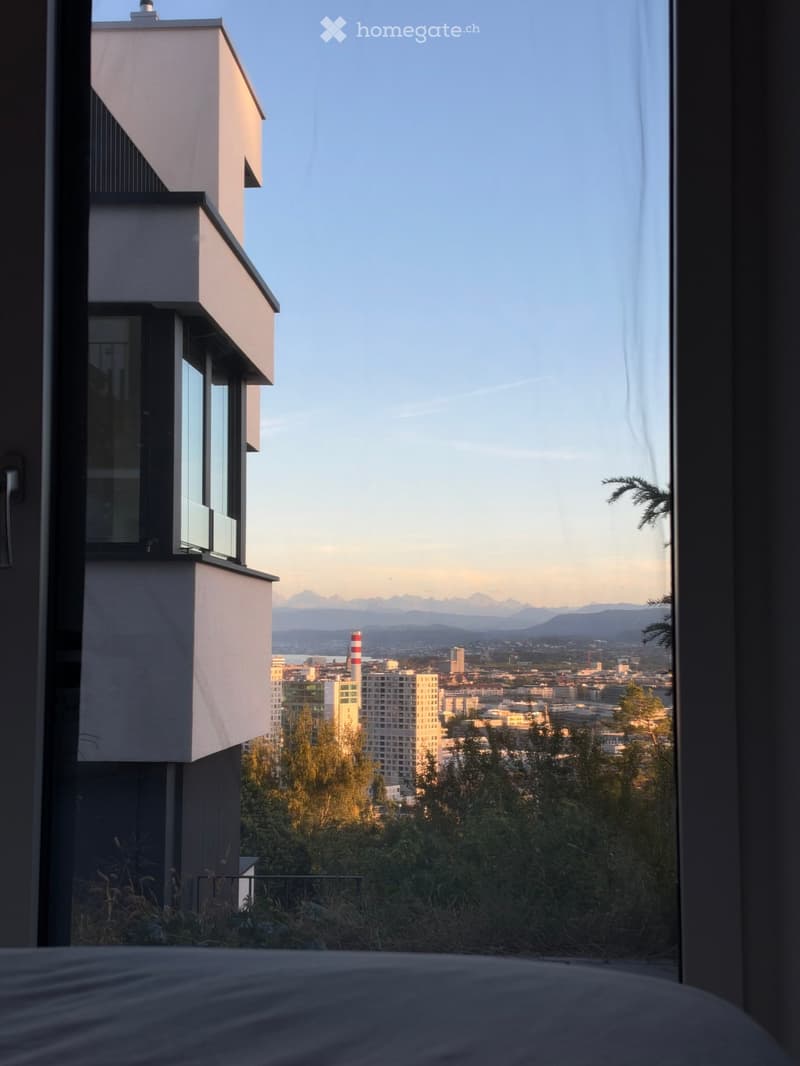 Bedroom with a large window offering a panoramic cityscape view, including buildings, trees, and distant mountains.