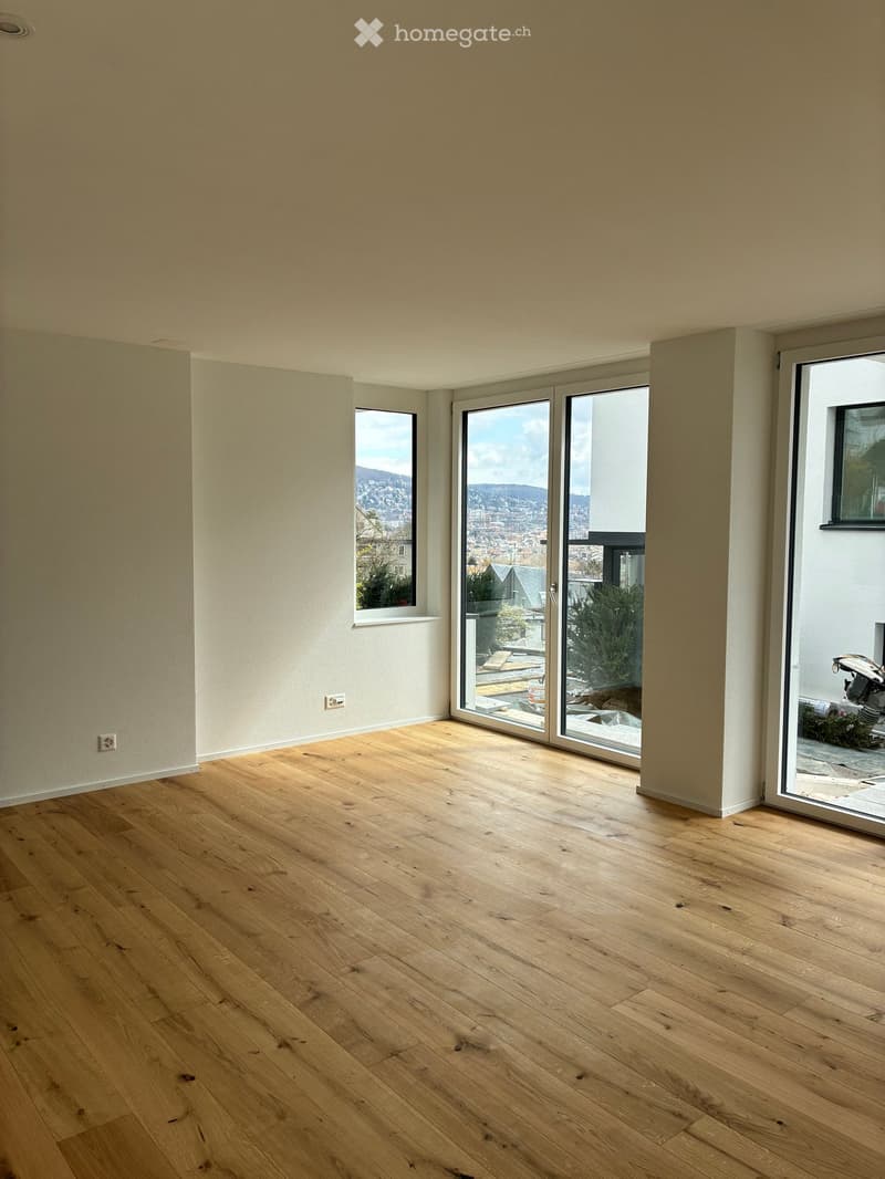 Empty room with light wooden flooring, white walls, and large windows offering a view of the city and surrounding landscape.