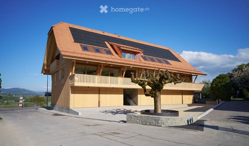 Modern two-story house with a wooden facade, solar panels on the roof, large windows, and a balcony overlooking the surrounding landscape.