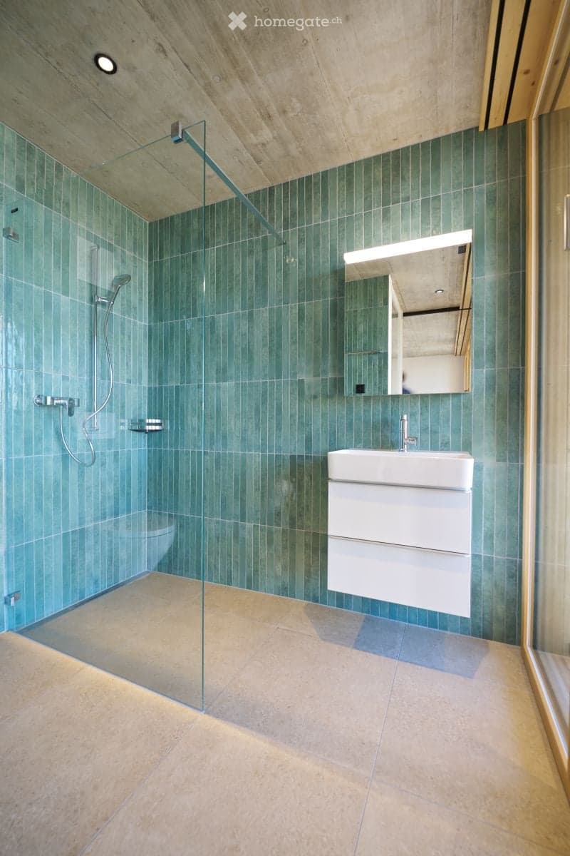 Modern bathroom with green tiled walls, a glass-enclosed shower, a white sink with a mirror, and a toilet.