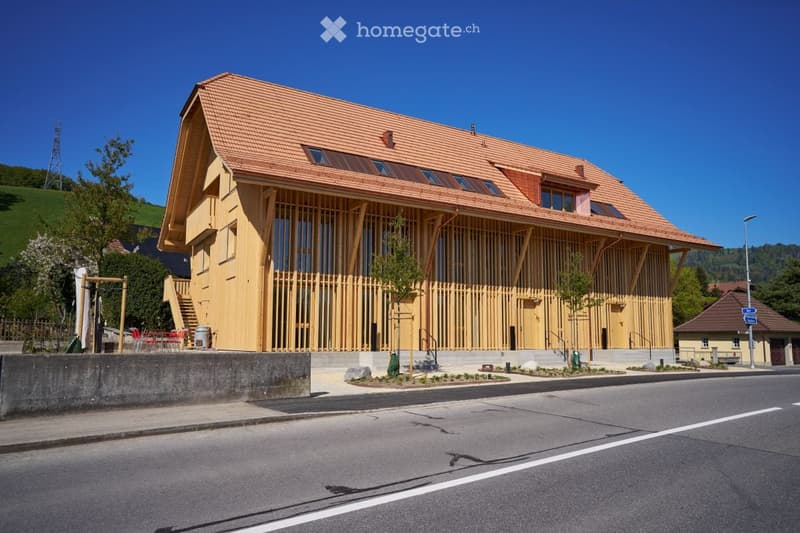 Modern wooden building with a red-tiled roof, large windows, and a raised ground floor, situated on a street with a small landscaped area in front.